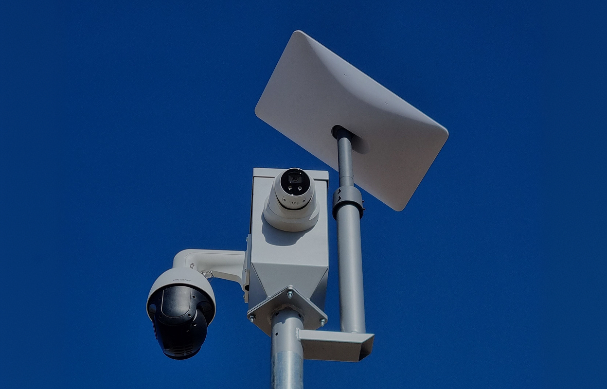 Two surveillance cameras mounted on a gray pole against a clear blue sky.